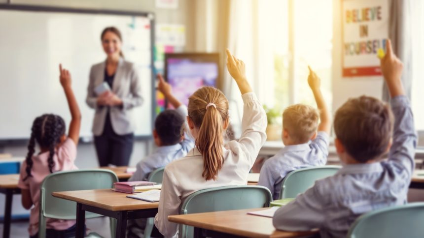 Modern classroom interior with students raising hands seen from behind and smiling happy teacher in background, concept of education and school life When the Money Doesn’t Follow the Students: America’s Education Funding Crisis