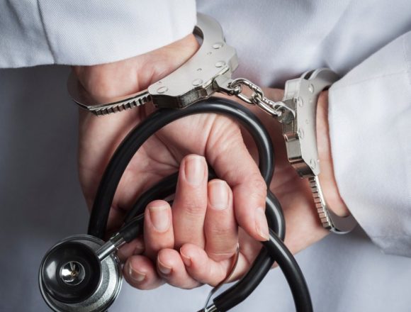Handcuffed hands behind a white lab coat holding a stethoscope, illustrating health care fraud enforcement